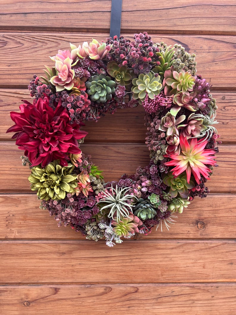 May include: A green and burgundy succulent wreath with a variety of succulents and burgundy flowers. The wreath is hanging on a wooden door.