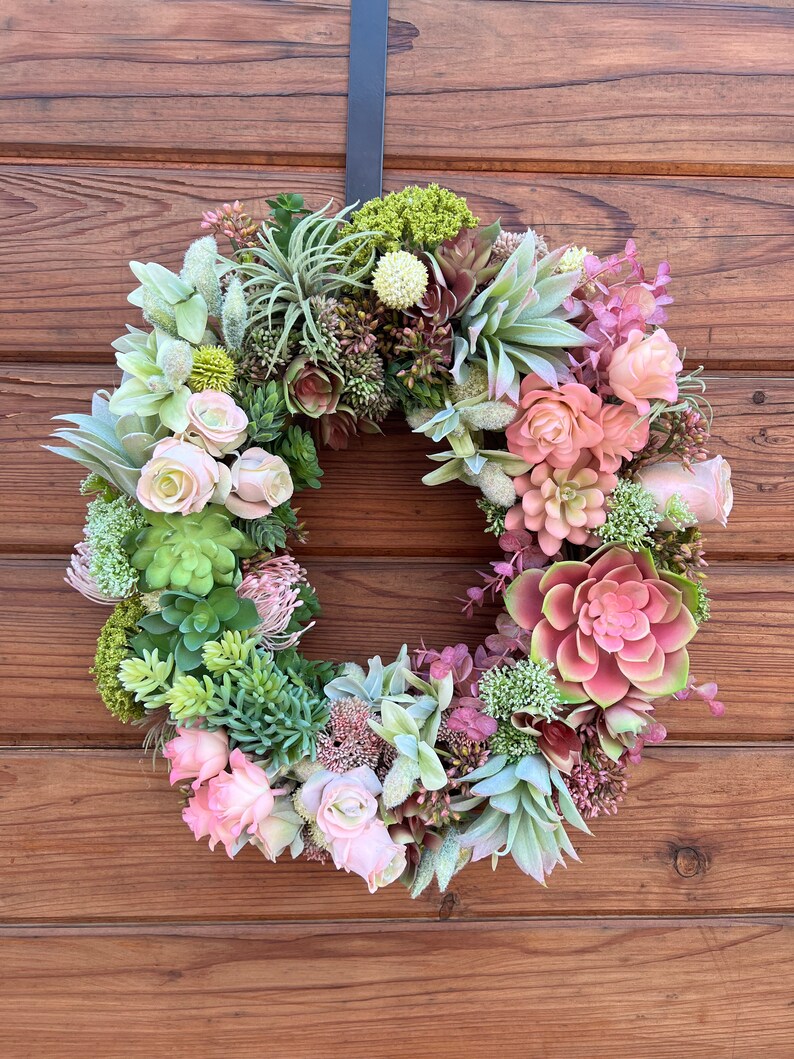 May include: A green and pink succulent wreath with pink roses hanging on a wooden door.