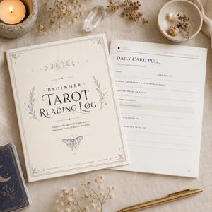 May include: A cream-colored Beginner Tarot Reading Log book with a moth illustration and moon phases on the cover. Beside it lies an open page titled Daily Card Pull with lines for date, card pulled, and notes. A gold pen and tarot card are nearby.