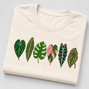 May include: A cream-coloured t-shirt featuring an embroidered row of six distinct tropical plant leaves. The design includes various shades of green, with one leaf displaying pink and green variegation and another featuring white polka dots.