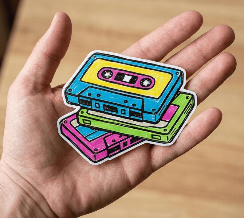 May include: A sticker featuring a stack of three illustrated cassette tapes in bright colors. The top tape is blue with a yellow center, the middle tape is green, and the bottom tape is pink. Each cassette has black outlines and details.