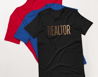 May include: Black t-shirt with the word "REALTOR" printed in gold on the front. The shirt is folded and displayed on top of a blue t-shirt and a red t-shirt.