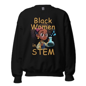 Puede incluir: Sudadera negra con un gráfico de una niña negra con gafas y sosteniendo un vaso de precipitados. El texto "Black Women in STEM" está impreso en amarillo en la sudadera.