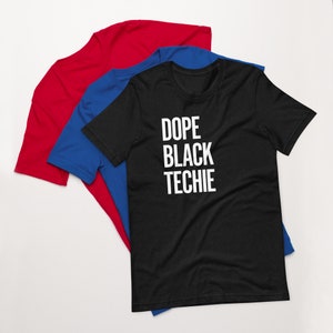 Pode incluir: Uma camiseta preta com o texto "Dope Black Techie" em letras brancas. A camiseta está dobrada e empilhada em cima de uma camiseta azul e uma camiseta vermelha.