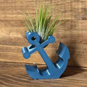 May include: A blue anchor-shaped planter holding a green air plant. The planter is made of a 3D-printed material and sits on a wooden surface. The anchor design features a classic shape with a ring at the top for the plant.