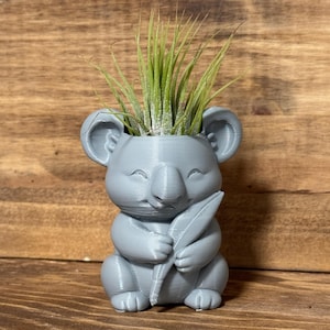 May include: A gray koala-shaped planter holding a leaf, with a green plant inside. The koala has a smiling expression and is made of a 3D-printed material. The planter is sitting on a wooden surface.