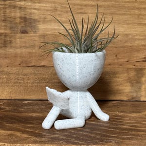 May include: A speckled white planter in the shape of a person, holding a book and with a plant inside. The planter has a round head and is sitting with its legs extended. The plant has long, thin green leaves.