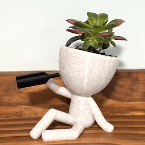 May include: White ceramic planter shaped like a person sitting down with a small black bottle in one hand. The planter has a speckled finish and is holding a green succulent plant.