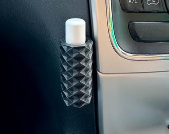 May include: A black, geometric-patterned car air vent clip holding a small white-capped bottle. The clip has a textured, diamond-like design. The bottle is cylindrical and appears to be a lip balm or similar product.
