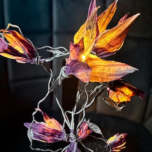 Könnte beinhalten: Eine dekorative Blumenlampe mit beleuchteten Blütenblättern in Orange-, Gelb- und Lilatönen. Die zarten Blütenblätter der Lampe sind auf dünnen, silberfarbenen Drahtstielen angeordnet und schaffen eine künstlerische und elegante Leuchte.
