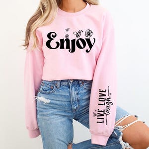 Pode incluir: Uma camisa de moletom rosa com a palavra "Enjoy" impressa em preto na frente. As mangas t&ecirc;m o texto "Live Love Laugh" impresso em preto.
