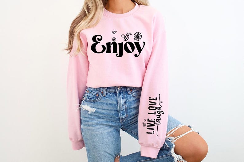 Pode incluir: Uma camisa de moletom rosa com a palavra "Enjoy" impressa em preto na frente. As mangas t&ecirc;m o texto "Live Love Laugh" impresso em preto.