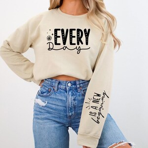 Pode incluir: Uma camisa de moletom bege com o texto "EVERY Day" na frente e "IS A NEW beginning" na manga. A modelo est&aacute; usando jeans azul.