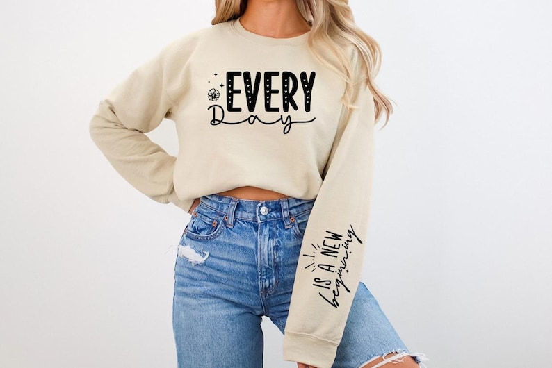 Pode incluir: Uma camisa de moletom bege com o texto "EVERY Day" na frente e "IS A NEW beginning" na manga. A modelo est&aacute; usando jeans azul.