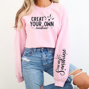 Pode incluir: Camisola cor-de-rosa com o texto "Crie o seu pr&oacute;prio sol" na frente e "Voc&ecirc; &eacute; o sol" numa manga.
