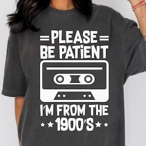 Op de afbeelding: Donkergrijs t-shirt met een witte afbeelding van een cassettebandje en de tekst "PLEASE BE PATIENT I'M FROM THE 1900'S". Het shirt heeft korte mouwen en een ronde hals.