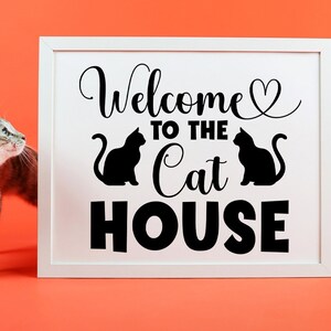 May include: White framed sign with black text that reads "Welcome to the Cat House" with two black cat silhouettes. A gray tabby cat is looking at the sign.