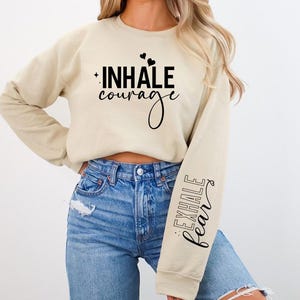 Pode incluir: Uma camisa de moletom bege com o texto "INHALE courage" na frente e "EXHALE fear" na manga.