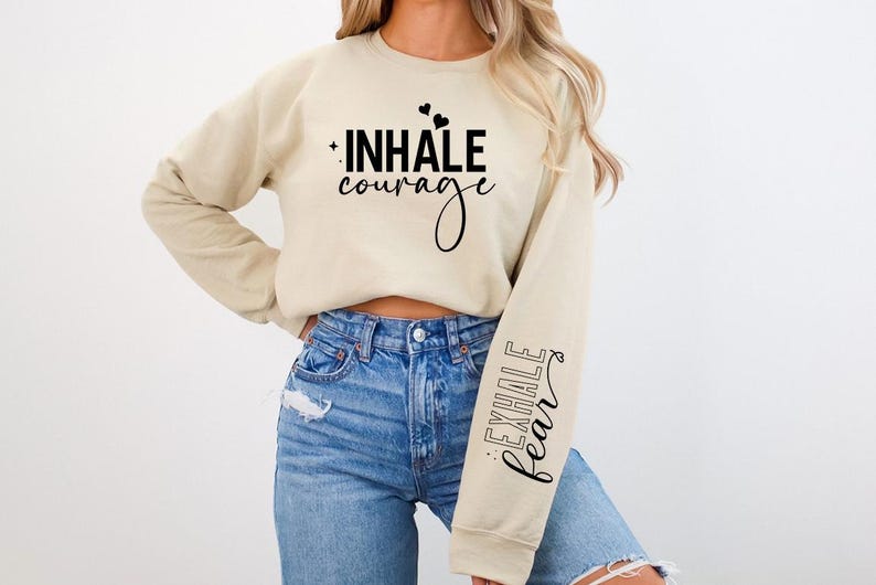 Pode incluir: Uma camisa de moletom bege com o texto "INHALE courage" na frente e "EXHALE fear" na manga.