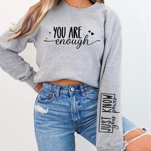 Pode incluir: Camisola de moletom cinza com o texto "You are enough" na frente e "Just know your power" na manga.