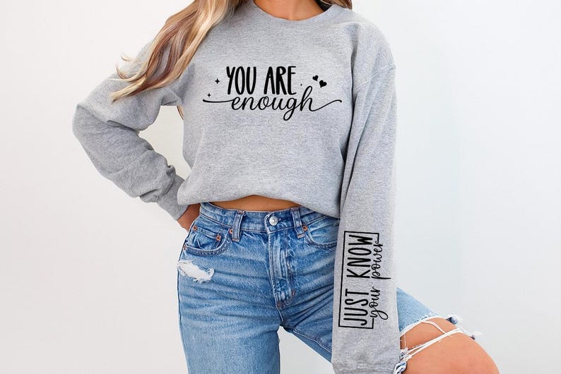 Pode incluir: Camisola de moletom cinza com o texto "You are enough" na frente e "Just know your power" na manga.