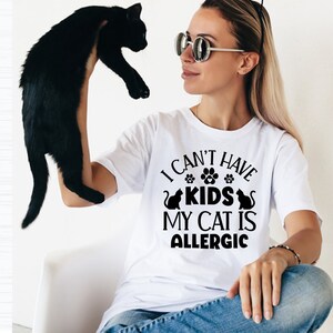 May include: A woman wearing a white t-shirt with the text "I can't have kids my cat is allergic" in black lettering. She is holding a black cat in her left hand.
