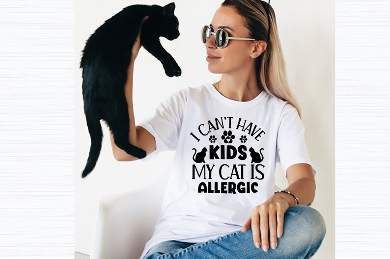 May include: A woman wearing a white t-shirt with the text "I can't have kids my cat is allergic" in black lettering. She is holding a black cat in her left hand.