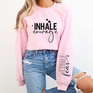 Pode incluir: Camisola cor-de-rosa com o texto "INHALE courage" na frente e "EXHALE fear" na manga.