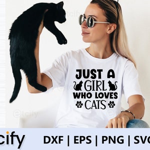 May include: A white t-shirt with the text "Just a girl who loves cats" in black lettering. The text is surrounded by two black cat silhouettes and two paw prints. A woman is holding a black cat in her left hand.