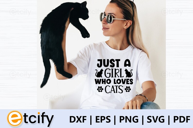 May include: A white t-shirt with the text "Just a girl who loves cats" in black lettering. The text is surrounded by two black cat silhouettes and two paw prints. A woman is holding a black cat in her left hand.