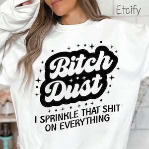 May include: White sweatshirt with the text "Bitch Dust" in a bold, black, stylized font, surrounded by stars and dots. Below the main text is the phrase "I SPRINKLE THAT SHIT ON EVERYTHING" in a sans-serif font.
