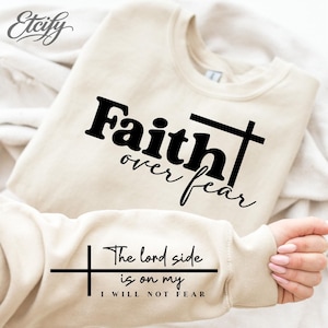 May include: A cream-colored sweatshirt with the words "Faith over fear" and a cross graphic in black. The sleeve displays the text "The lord side is on my, I will not fear" in black. The Etsy logo is in the upper left corner.