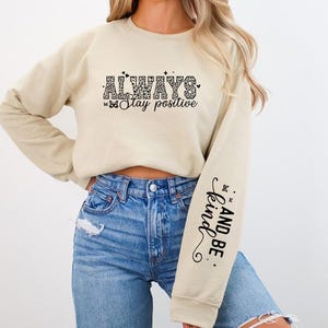 Pode incluir: Uma camisa de moletom bege com o texto "ALWAYS Stay positive" na frente e "AND BE kind." na manga.