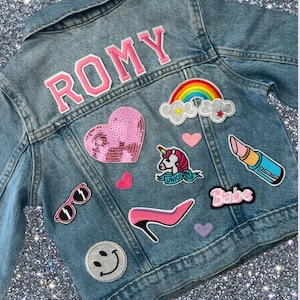 Pode incluir: Casaco de ganga azul claro com o nome "ROMY" em letras cor-de-rosa. O casaco é decorado com aplicações, incluindo um coração com lantejoulas, um arco-íris, um unicórnio, um batom, um salto alto, óculos de sol, um smiley e a palavra "Babe".