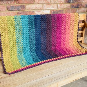 Puede incluir: Una manta de ganchillo colorida con un degradado de verde, azul, morado y naranja. La manta tiene un borde de ganchillo marr&oacute;n.