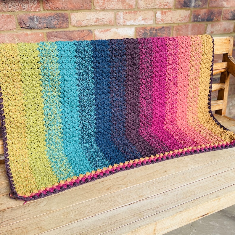 Puede incluir: Una manta de ganchillo colorida con un degradado de verde, azul, morado y naranja. La manta tiene un borde de ganchillo marr&oacute;n.