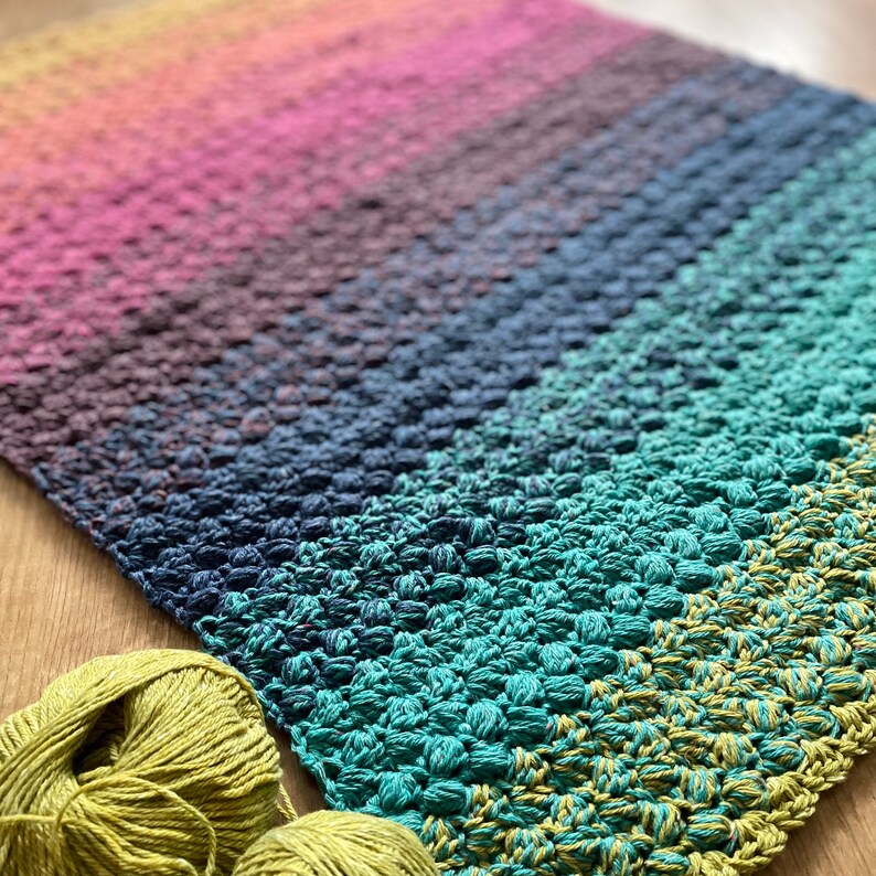 Puede incluir: Una manta de crochet colorida con un degradado de rosa, morado, azul, verde y amarillo. La manta est&aacute; hecha con un punto texturizado que crea una superficie abultada.
