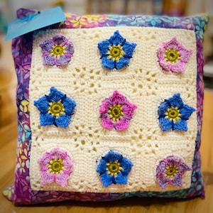 Könnte beinhalten: Ein weißes gehäkeltes Kissen mit Blumenmuster. Das Kissen ist aus weißem Garn gefertigt und zeigt ein sich wiederholendes Muster aus blauen, rosa und grünen Blumen. Das Kissen ist von einem lila und weißen Stoffrand umgeben.