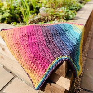 Puede incluir: Una manta de crochet colorida con un degradado de amarillo, naranja, rosa, morado, azul y verde. La manta est&aacute; doblada y drapeada sobre una superficie de madera.