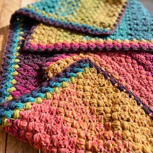 Puede incluir: Primer plano de una manta de ganchillo con un patr&oacute;n multicolor. La manta est&aacute; hecha con diferentes tonos de rosa, amarillo, azul y verde. La manta est&aacute; doblada por la mitad, mostrando el intrincado patr&oacute;n de puntadas.