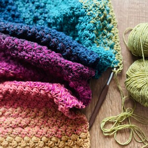 Puede incluir: Una manta de ganchillo multicolor con un degradado de azul, morado, rosa y naranja. La manta est&aacute; doblada por la mitad y se est&aacute; trabajando con un gancho de ganchillo y lana.