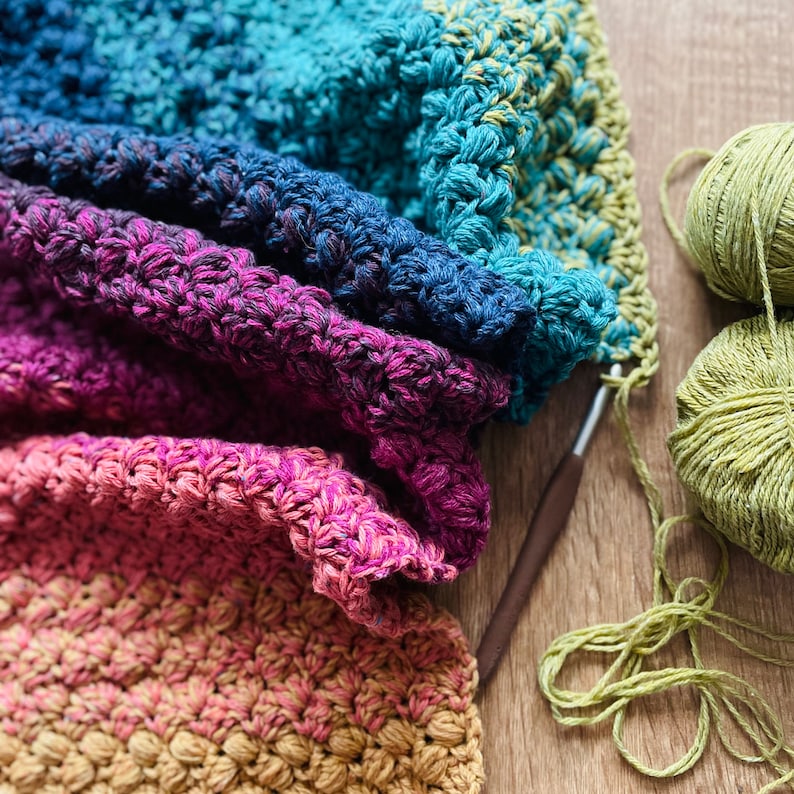 Puede incluir: Una manta de ganchillo multicolor con un degradado de azul, morado, rosa y naranja. La manta est&aacute; doblada por la mitad y se est&aacute; trabajando con un gancho de ganchillo y lana.