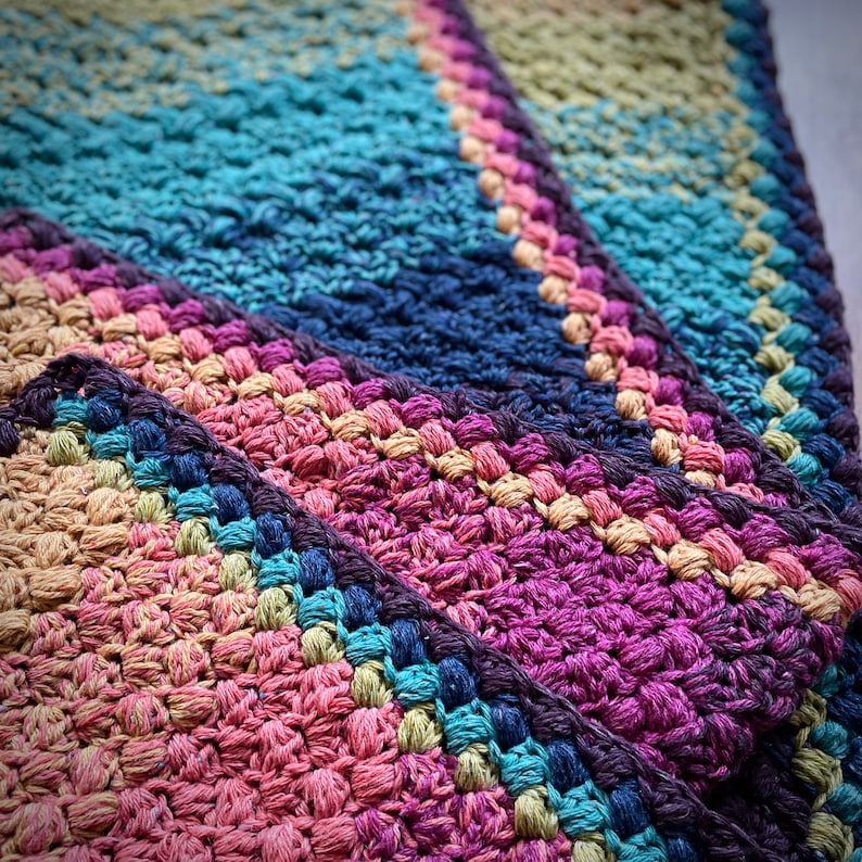 Puede incluir: Alfombra de crochet multicolor con un dise&ntilde;o en capas. La alfombra est&aacute; hecha de hilo grueso y tiene una superficie texturizada. Los colores son una mezcla de azules, morados y verdes.