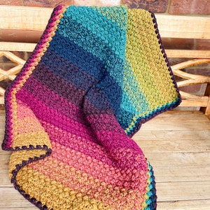 Puede incluir: Una manta de ganchillo colorida con un degradado de azul verdoso, azul, morado, rosa, naranja y amarillo. La manta tiene un borde marr&oacute;n oscuro.