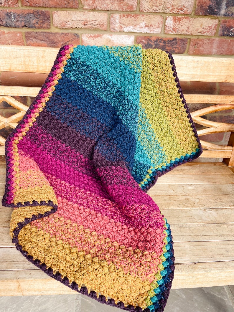 Puede incluir: Una manta de ganchillo colorida con un degradado de azul verdoso, azul, morado, rosa, naranja y amarillo. La manta tiene un borde marr&oacute;n oscuro.