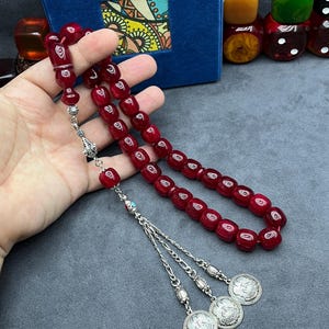 May include: A red prayer bead rosary with silver accents and three decorative coin pendants. The beads are a deep red color, and the silver components add contrast. The rosary is held in a hand, with a blue box and dice in the background.