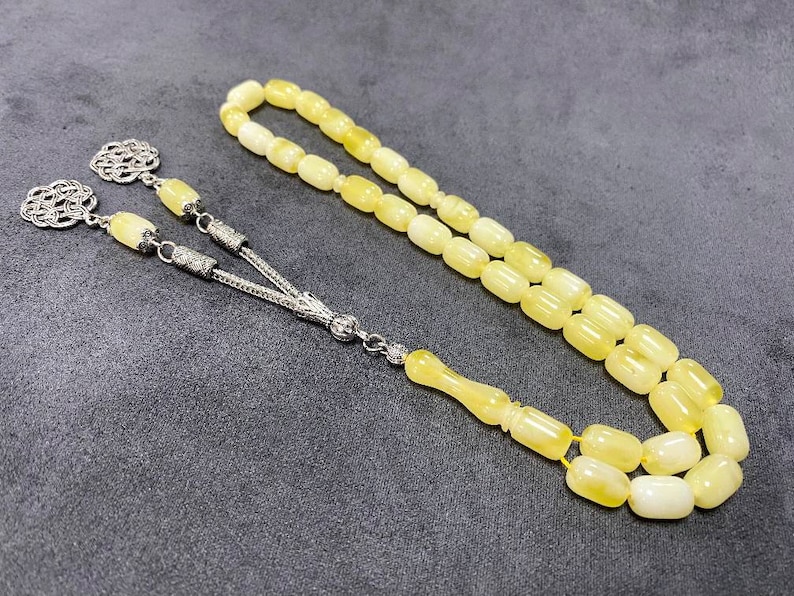 Puede incluir: Un collar de cuentas de oraci&oacute;n de &aacute;mbar amarillo con detalles plateados. Las cuentas son de forma ovalada y el collar tiene un borla.