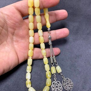Puede incluir: Un collar de cuentas de oraci&oacute;n de &aacute;mbar amarillo con detalles plateados. Las cuentas son de forma ovalada y el collar tiene un borla con un dise&ntilde;o de nudo.
