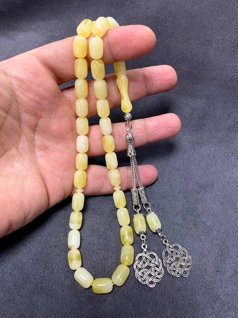 Puede incluir: Un collar de cuentas de oraci&oacute;n de &aacute;mbar amarillo con detalles plateados. Las cuentas son de forma ovalada y el collar tiene un borla con un dise&ntilde;o de nudo.