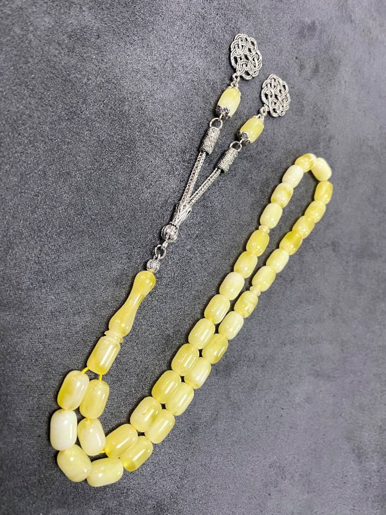 Puede incluir: Un collar de cuentas de oraci&oacute;n de &aacute;mbar amarillo con detalles plateados. Las cuentas son de forma ovalada y el collar tiene un borla.